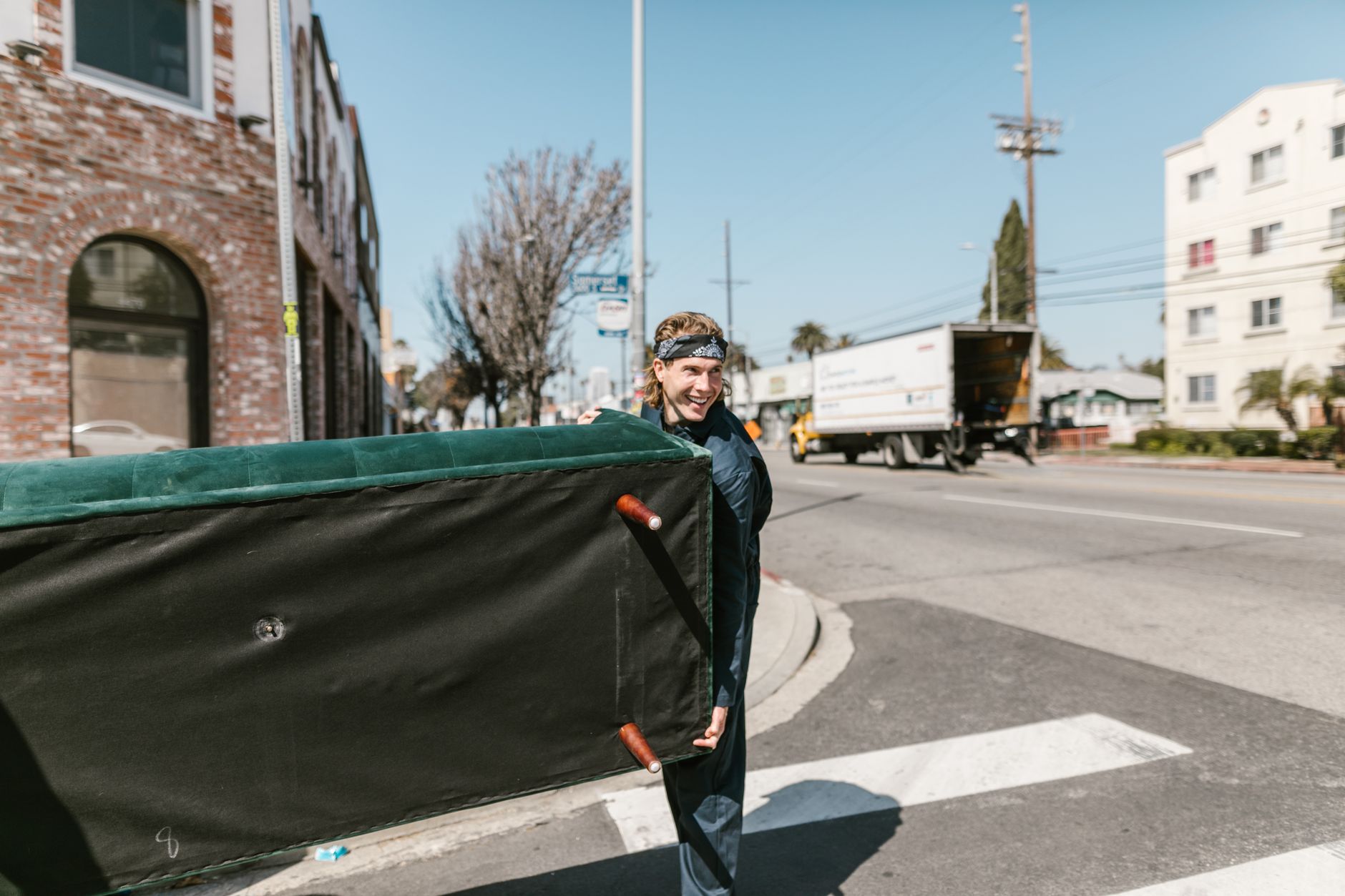 Man carrying a couch across a sunny street during a move, similar to a couch removal near me pickup in Vero Beach